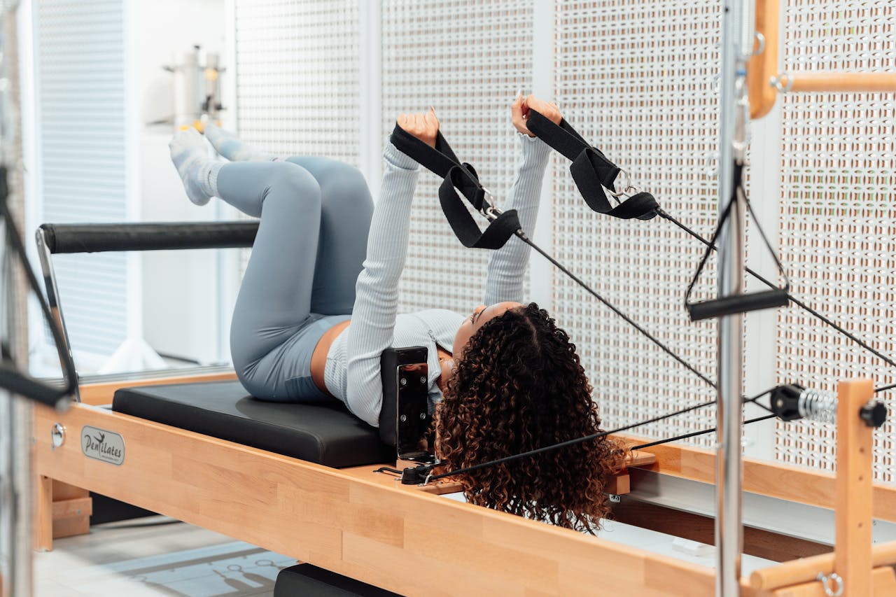 A woman wearing a gray tracksuit exercises on a Pilates reformer in a modern studio setting.